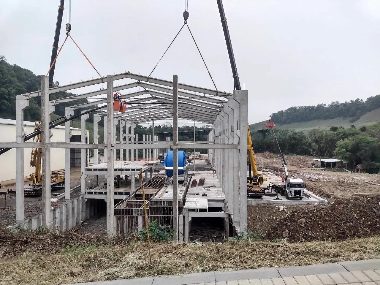 Montagem de estrutura pré-moldada de galpão com dois guindastes, pilares e vigas de cobertura
